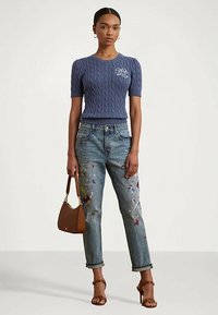 Γυναίκα φοράει ένα μπλε κοντομάνικο πουλόβερ με πλέξη κασμίρ, τζιν που είναι γεμάτα μπογιές, καφέ γόβες και κρατάει μια καφέ τσάντα.