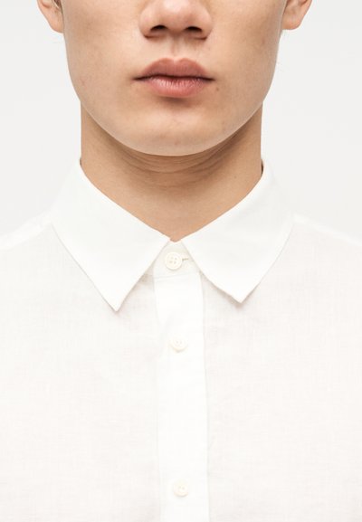 Κοντινή άποψη του κάτω μέρους του προσώπου και του λαιμού ενός ατόμου που φοράει λευκό πουκάμισο με κουμπιά και μυτερό γιακά, πάνω σε απλό φόντο.
