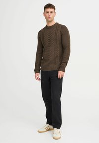 Brauner gestrickter Pullover mit Zopfmuster, Rundhalsausschnitt und gerippten Bündchen, kombiniert mit schwarzen Jeans und weißen Sneakers mit braunen Streifen.