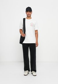 T-shirt de algodão branca com um logotipo colorido, calças de veludo cotelê pretas, e um boné preto, segurando uma mala de ombro preta, com sapatilhas bege, em um fundo neutro.