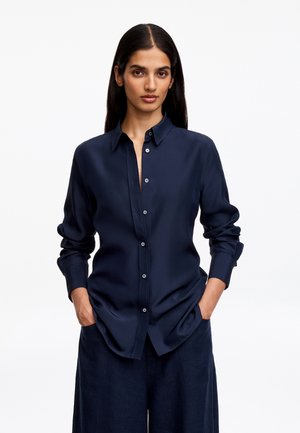 Femme aux longs cheveux foncés porte une chemise bleu foncé boutonnée et un pantalon foncé, debout les mains dans les poches devant un fond blanc uni.