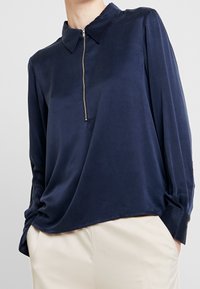 Blusa a maniche lunghe blu navy in tessuto lucido con colletto con zip. Presenta un morbido drappeggio e polsini con bottoni, abbinata a pantaloni crema.