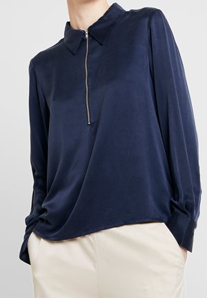 Marineblaue Bluse mit langen Ärmeln aus glattem Stoff und einem Reißverschlusskragen. Sie verfügt über sanfte Drapierungen und Knopfmanschetten und wird mit cremefarbenen Hosen kombiniert.