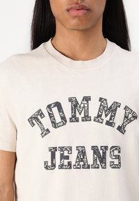 Šviesiai smėlio spalvos medvilninė marškinėliai su trumpomis rankovėmis, ant kurių ryškiai juodai atspausdintas tekstas "TOMMY JEANS" dekoratyviu šriftu.