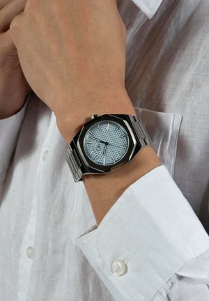 Orologio in acciaio inox argentato con cassa ottagonale, quadrante texturizzato blu chiaro e indici delle ore argentati, indossato su un polso con una camicia bianca.