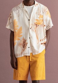 Man in een crème kortemouwen overhemd met een schelpen- en koraalprint, een witte onderhemd, en felgele broek met trekkoord.