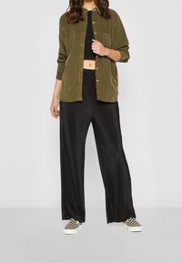 Camicia button-up in corduroy verde oliva sopra un top corto nero, abbinata a pantaloni neri a pieghe e larghi e scarpe slip-on a scacchi.