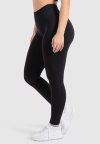 Leggings neri con vita alta, texture liscia e design aderente, abbinati a scarpe da ginnastica bianche con tomaia in mesh.