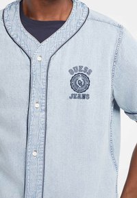 Lichtblauwe denim knoop-up overhemd met textuurstof, contrasterende donkerblauwe afwerking en een geborduurd logo met de tekst "GUESS JEANS."