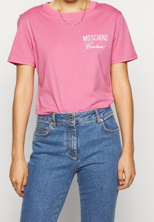Vrouw draagt een roze Moschino Couture T-shirt, gestopt in een hooggetailleerde blauwe spijkerbroek, met een gouden ketting.