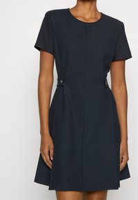 Mörkblå klänning med kort ärm och utsvängt kjol, med sidoknappar och ett slätt, texturerat tyg som framhäver en figursydd silhuett.
