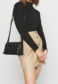 Schwarzes Langarmoberteil, beigefarbenen, hochgeschnittenen Rock mit sichtbaren Knöpfen, und eine kleine schwarze gesteppte Handtasche mit Schultergurt.