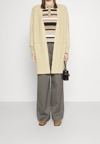 Cardigan beige e morbido con tasche laterali, indossato sopra una polo a righe nei toni del marrone, nero e crema. Pantaloni larghi scuri e scarpe a fantasia.