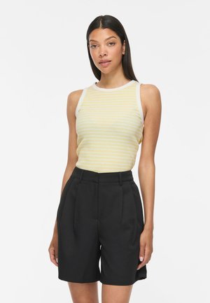 Femme aux longs cheveux foncés portant un top sans manches rayé jaune et blanc et un short noir taille haute ajusté sur un fond uni.