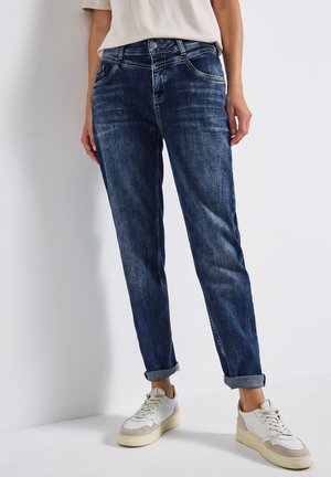 Jean en denim bleu foncé avec un délavage, comportant des poches avant, des poignets roulés et des détails de couture horizontale. Associé à des baskets blanches.