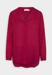 Blouse à manches longues, rouge foncé, avec boutons cachés sur le devant et col pointu, présentée sur un fond clair uni.