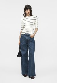 Woman wearing a white top with black horizontal stripes, wide-leg blue jeans, dark shoes, holding a black handbag, standing against a plain background.