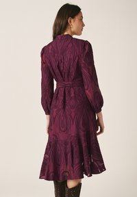 Frau trägt ein langärmeliges, knielanges lila gemustertes Kleid mit einem Stoffgürtel, steht seitlich vor einem einfarbigen Hintergrund.