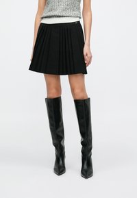 Zwarte geplooide rok met een nauwsluitende tailleband en gedetailleerde plooien. Gecombineerd met kniehoge zwarte leren laarzen.