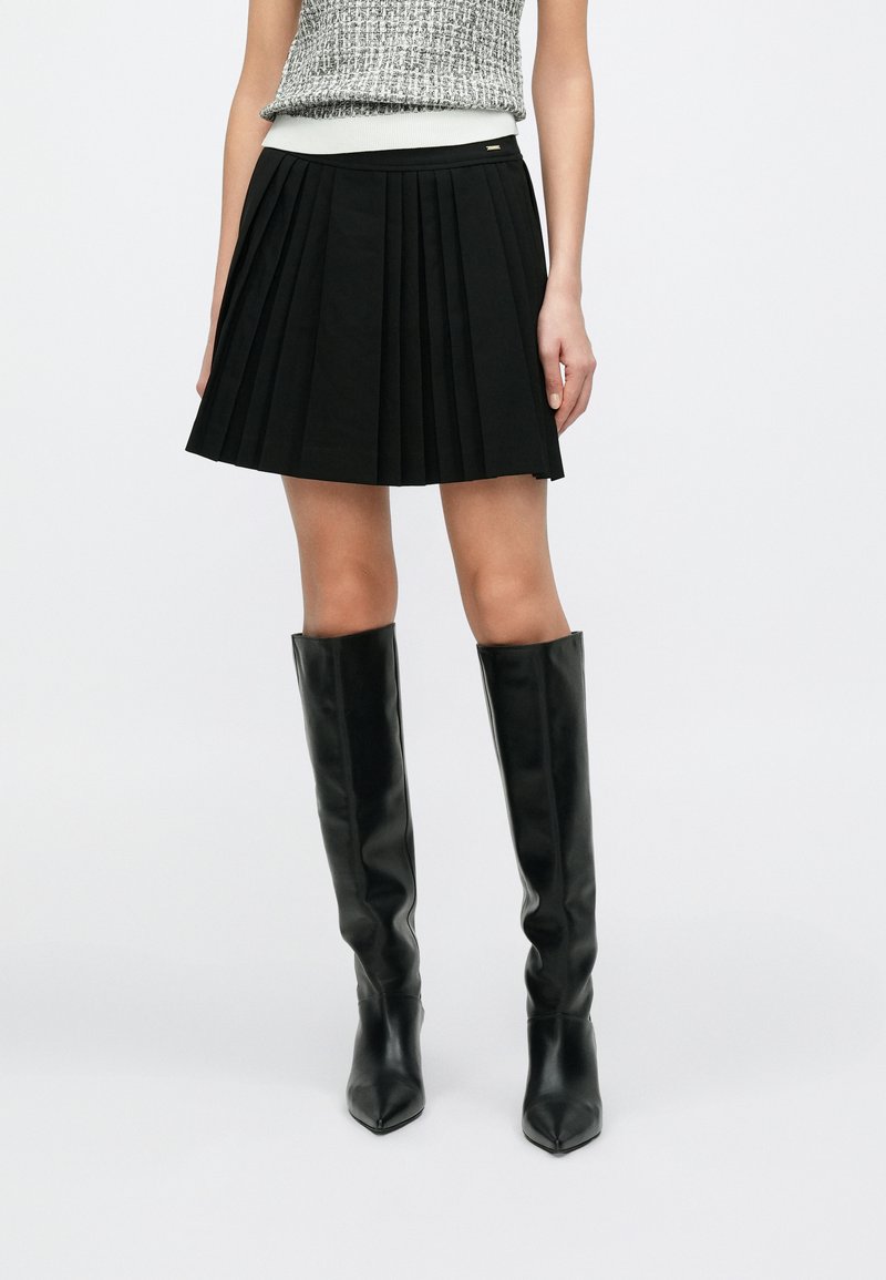 Zwarte geplooide rok met een nauwsluitende tailleband en gedetailleerde plooien. Gecombineerd met kniehoge zwarte leren laarzen.