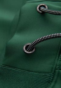 Mörkgrön tyg med en strukturfinish, med en svart och vit mönstrad dragsko som är trätt genom en metallöga med varumärke.