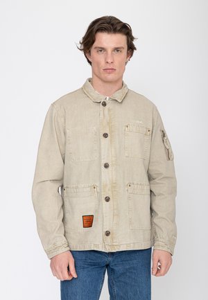 Jonge man die een beige jas met knopen en meerdere zakken draagt en een blauwe spijkerbroek, staand tegen een effen witte achtergrond.