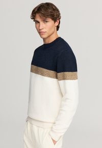 Młody mężczyzna stoi z rękami w kieszeniach, ubrany w sweter w kolorowe bloki w odcieniach granatowym, beżowym i białym, na jednolitym jasnym tle.