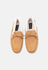 Et par mokkasiner i tan suede med syede detaljer, bundet sløjfer og hvid fleeceforing, mærket Pier One, på en hvid baggrund.