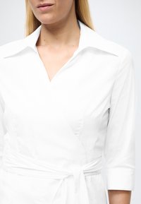 Vestito avvolgente bianco con scollo a collo, maniche a tre quarti e vita annodata. Il tessuto liscio e le linee pulite caratterizzano il design.