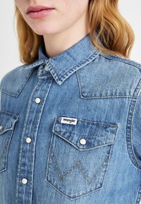 Ανοιχτό μπλε τζιν πουκάμισο με κλασικό γιακά, κουμπιά με σnap, δύο τσέπες στο στήθος και ετικέτα Wrangler. Διαθέτει ξεθωριασμένη υφή και αντίθετες ραφές.