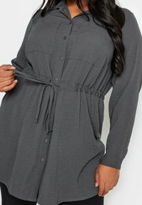 Grijze button-up shirt met lange mouwen en een trekkoord in de taille. Beschikt over twee voorkruiden en een gestructureerde stof.