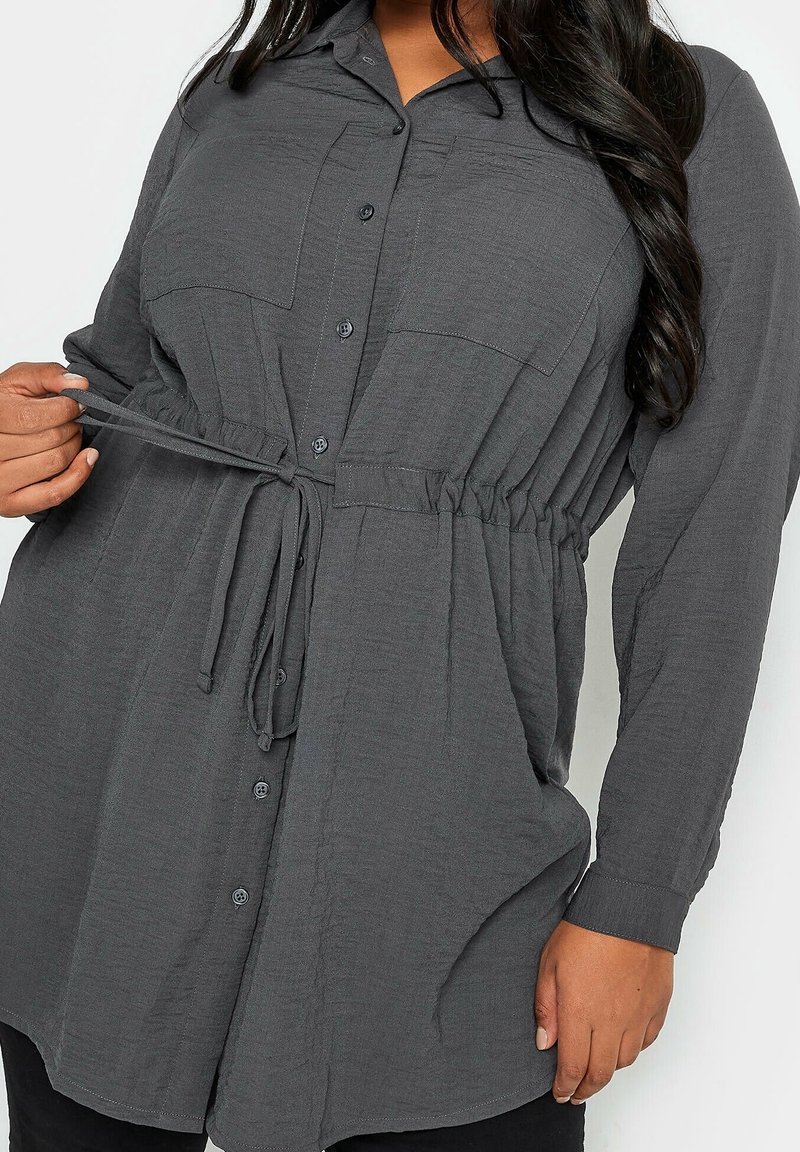 Grijze button-up shirt met lange mouwen en een trekkoord in de taille. Beschikt over twee voorkruiden en een gestructureerde stof.