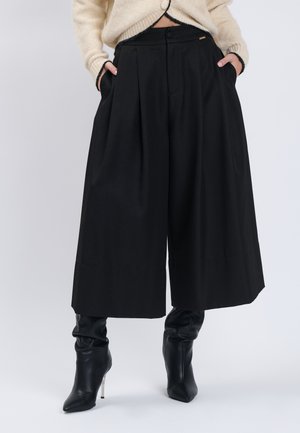 Person iført sorte culottebukser med vidde, sorte støvler med hæl og en beige cropped sweater med hænderne i lommerne.