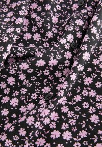 Tissu noir avec un petit motif floral rose clair et des boutons noirs, présenté avec de légers plis et un détail texturé en gros plan.