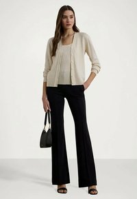 Moteris stovi vilkėdama smėlinį raštuotą kardiganą ir marškinėlius, juodas platformas, juodą flared kelnes ir laiko mažą juodą rankinę.