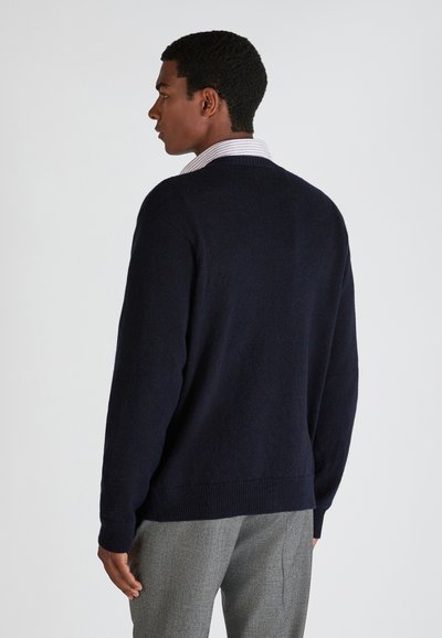 Pull en maille marine avec un ourlet côtelé et un col rond, porté par-dessus une chemise à col à rayures claires, vu de derrière.