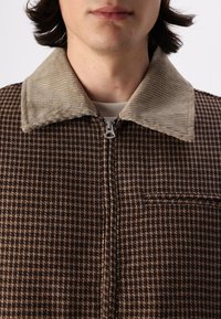 Chaqueta de cuadros marrón con un collar de pana beige, cremallera frontal y un bolsillo en el pecho. Tejido texturizado y detalles de diseño estructurados.