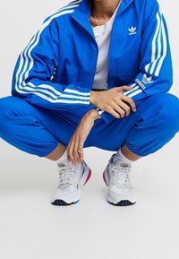 Blå Adidas träningsoverall med vita ränder, matchande joggers och grå sneakers med blå och rosa detaljer. I en knästående position mot en neutral bakgrund.