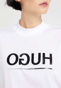 Camiseta de algodón blanca con un destacado logo impreso en negro "HUGO" y una línea horizontal debajo. Cuello alto, textura suave.