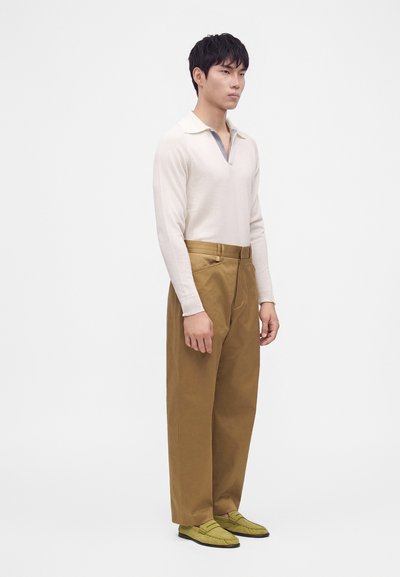 Homme debout portant une chemise blanche à manches longues, un pantalon marron à jambes larges et des mocassins vert clair sur fond blanc uni.