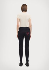 Black skinny jeans with back pockets featuring red embroidered text. Paired with a fitted cream top, highlighting a clean, simple design.