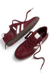 Paire de baskets bordeaux avec des bandes blanches sur les côtés, des semelles marron texturées et un laçage avant avec des lacets partiellement défaits sur fond blanc.