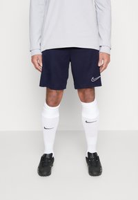 Bermudas atléticas en color navy con el logo blanco de Nike, combinadas con calcetas blancas hasta la rodilla que presentan el logo negro de Nike, y zapatillas deportivas negras.
