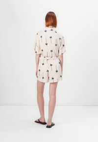 Rompers à manches courtes, couleur crème, avec un imprimé de palmiers noirs, taille élastique et shorts. Porté avec des sandales plates noires. Design minimaliste.