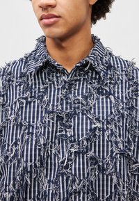 Svītrota tumši zilā krekls ar teksturētu, saplēstu dizainu. Tam ir pogu aizdare un klasiskā apkakle. Audums ietver zilas un baltas šķiedras.