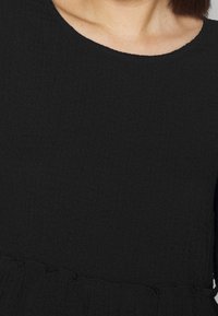 Schwarze Bluse aus strukturiertem Stoff, mit Rundhalsausschnitt und Rüschensaum. Einfarbig ohne Muster oder Verzierungen.