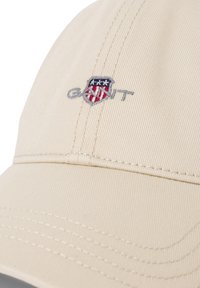 Beige katoenen pet met een geborduurd schildlogo in rood, wit en blauw, met gestikte details en een gebogen klep.