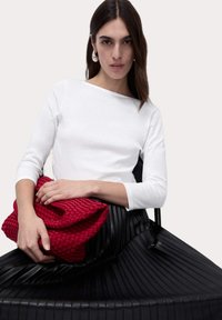 Red woven clutch bag held against a white long-sleeve top layered over a black pleated skirt, highlighting texture and color contrast.