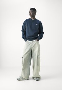 Felpa a girocollo navy, pantaloni cargo azzurri dal taglio loose fit, tessuto testurizzato, tasche laterali prominenti, abbinati a sneakers grigie.