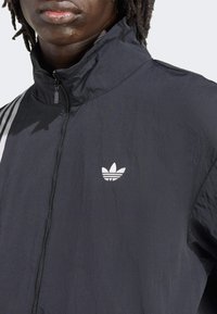 Black zip-up jacket with a high collar, featuring white Adidas logo and three white stripes on the sleeves. Lightweight fabric with textured appearance.